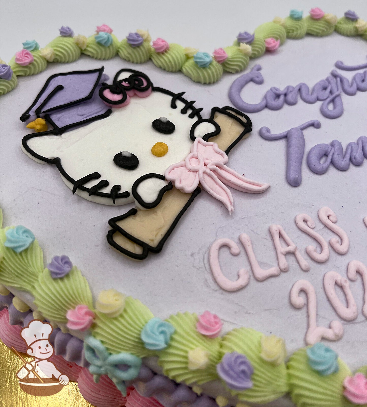 Heart-shaped cake with a pastel purple base, featuring a buttercream Hello Kitty face wearing a graduation cap and holding a diploma with a pink bow. Decorated with vintage-style pastel piping, small buttercream flowers, and buttercream bows along the edges.