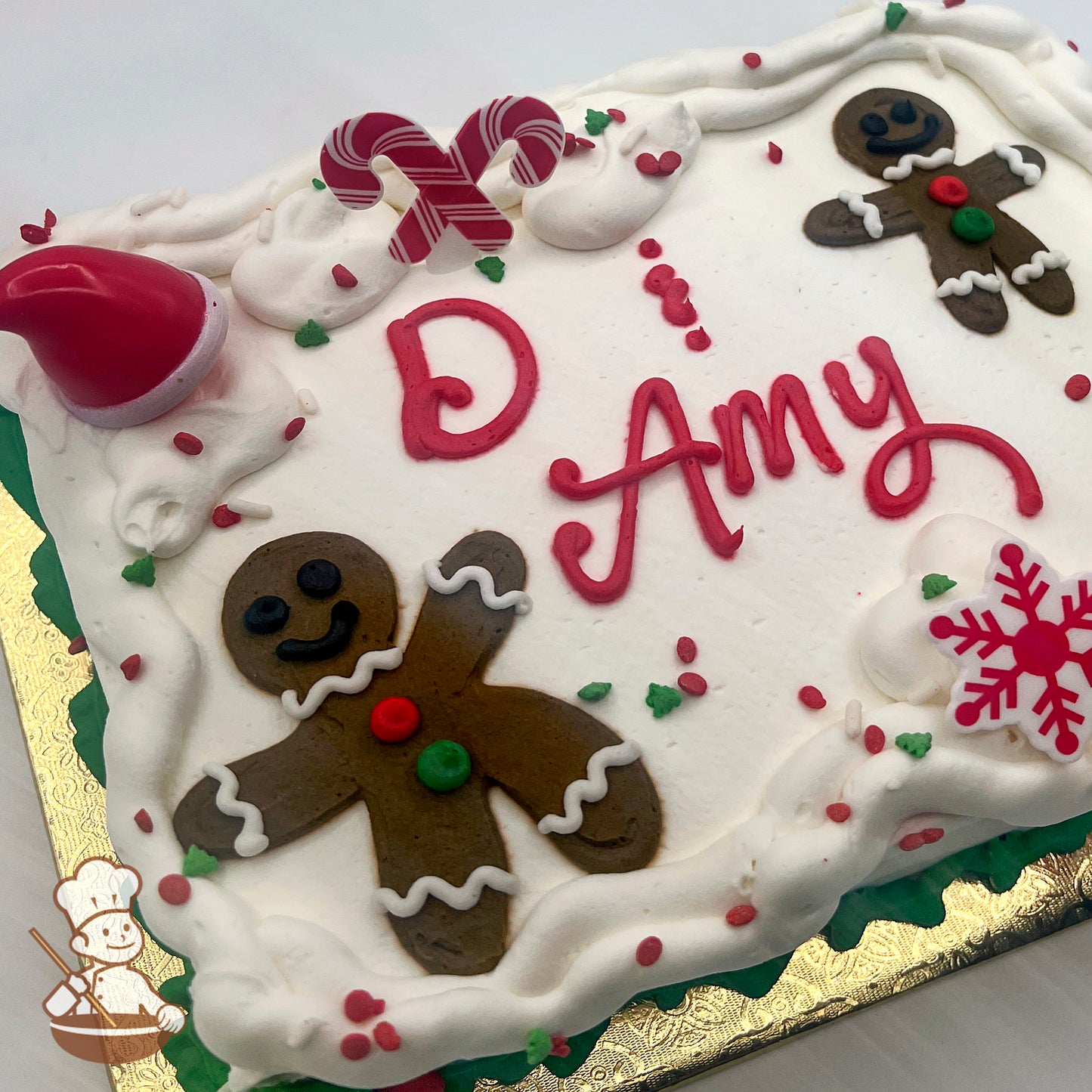 Sheet cake with white icing, decorated with plastic candy canes, a Santa hat, a red snowflake, buttercream gingerbread men and Christmas sprinkles.
