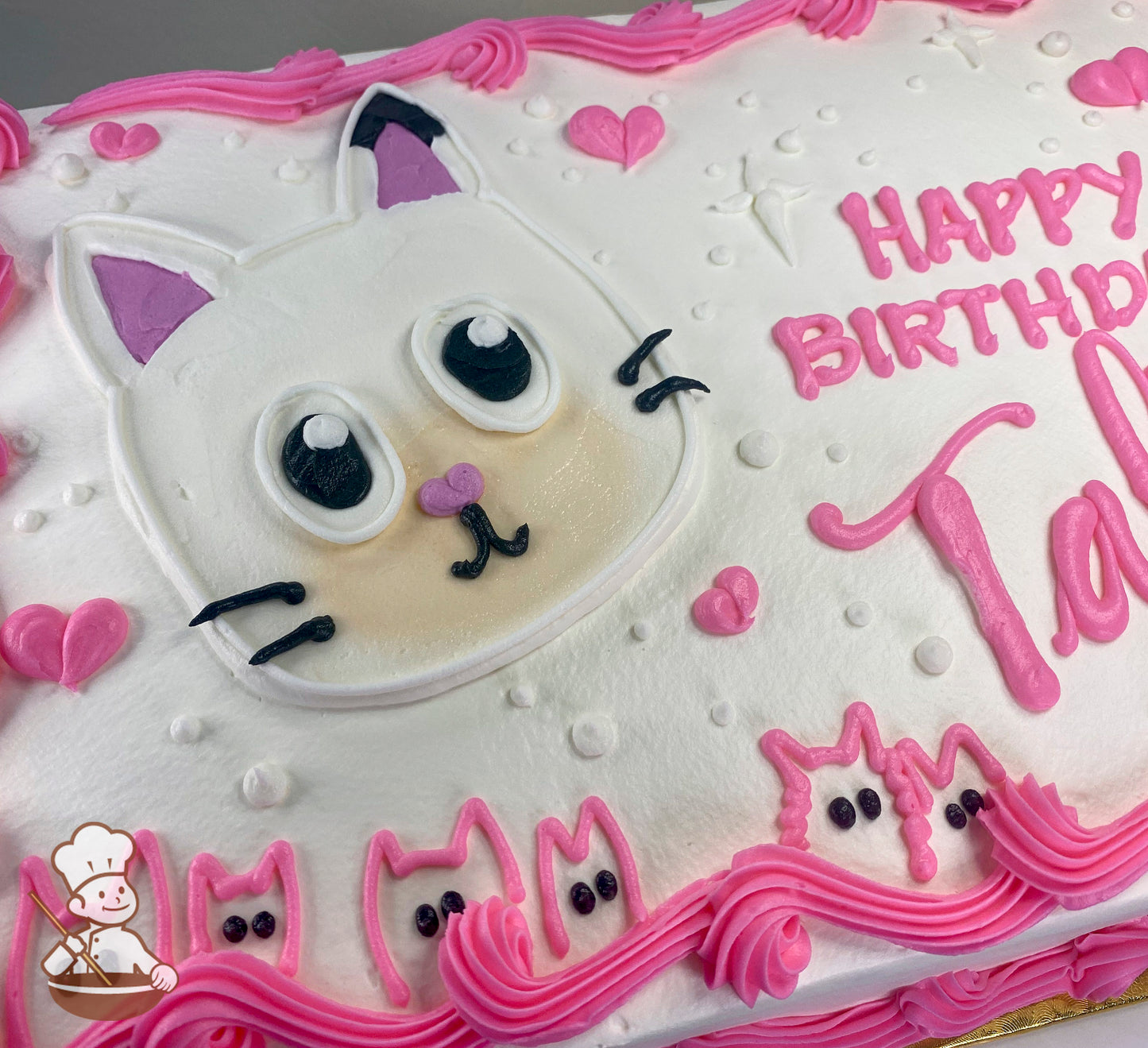 White sheet cake with a buttercream design of a Gabby's Dollhouse cat face, pink hearts, dots, and pink cat outlines along the border.