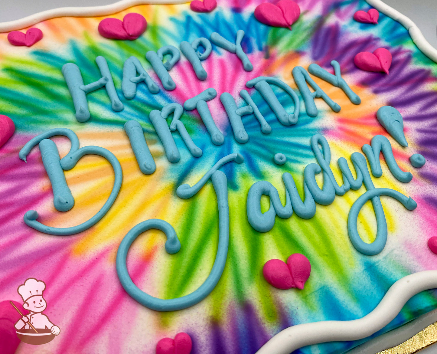 Colorful tie-dye sheet cake with vibrant swirls of pink, blue, yellow, green, and purple, decorated with pink buttercream hearts and a white border.