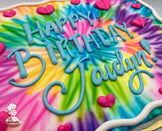 Colorful tie-dye sheet cake with vibrant swirls of pink, blue, yellow, green, and purple, decorated with pink buttercream hearts and a white border.