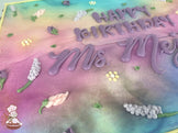 Sheet cake with a pastel gradient background of pink, purple, and blue, decorated with buttercream lavender flowers, small yellow blossoms, and green leaves.