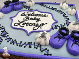Sheet cake with light gray icing, purple border, black stitch designs, white pumpkins with gray stems, and purple baby booties with black laces.