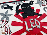 A sheet cake with buttercream decorations featuring a black ninja holding a sword, red sunburst design, gray shuriken's, and nun chucks.