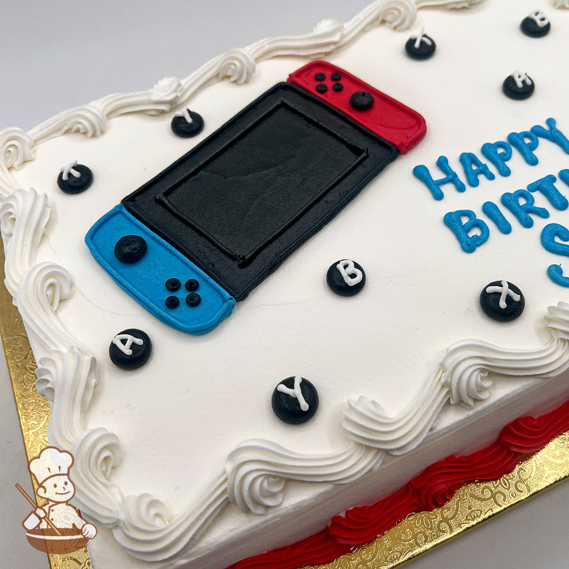 A sheet cake with white frosting, decorated with a Nintendo Switch design in the center. The Switch features red and blue Joy-Con controllers and a black screen. Black circular buttons with white letters "A," "B," "X," and "Y" are scattered around the cake. The edges are adorned with white swirl trims, and the base has a red buttercream border.