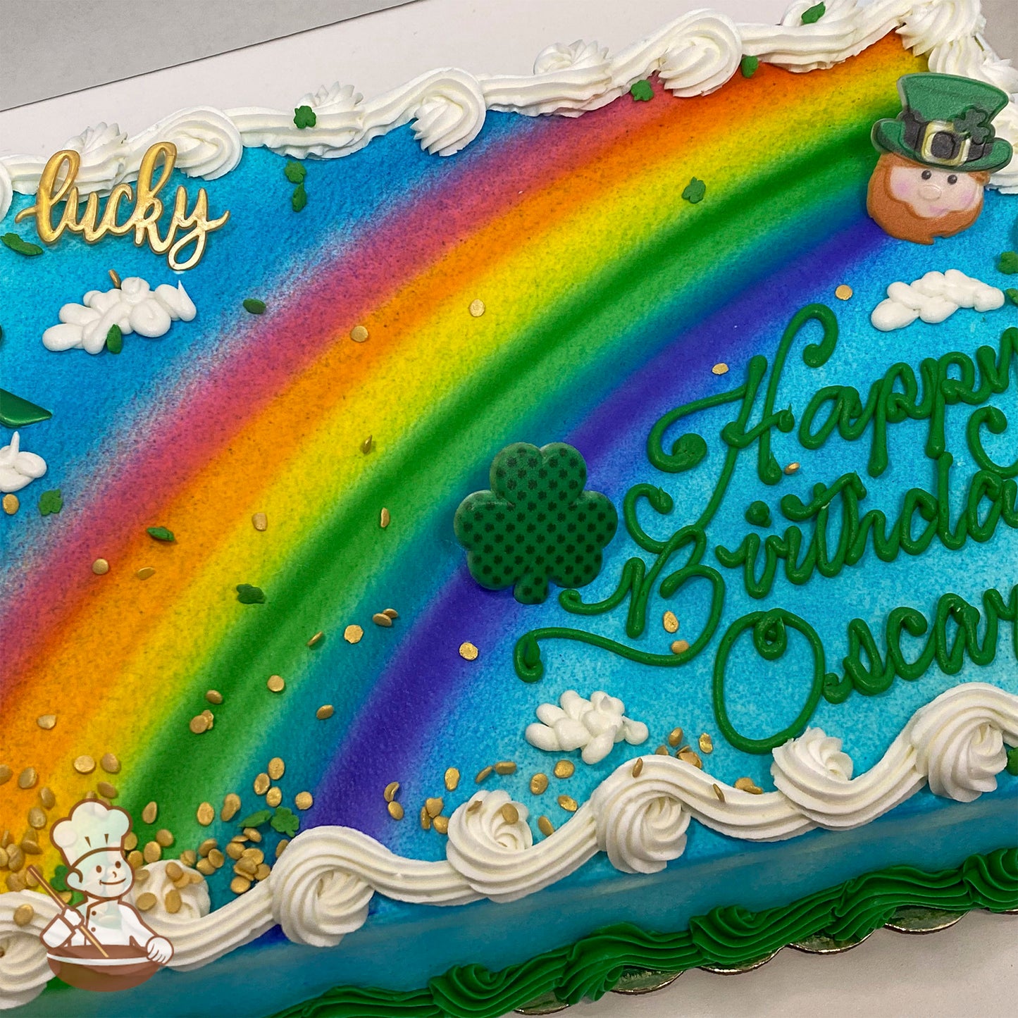 Sheet cake with a colorful rainbow design, white clouds, gold sprinkles, a plastic leprechaun face, shamrock, and piped buttercream borders.