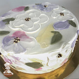 Round cake with white frosting, decorated with pastel painted flowers in pink, purple, and yellow, and green leaves.