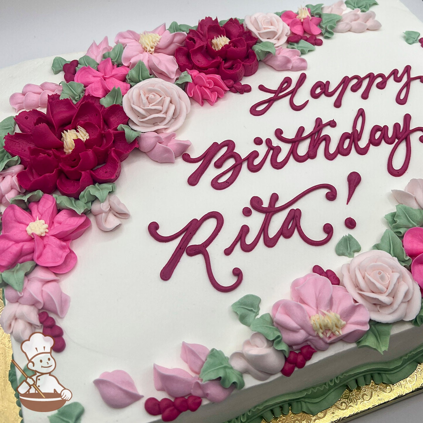 White sheet cake with buttercream decorations of pink and red peonies, roses, and assorted florals and accented with green leaves.