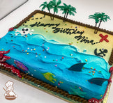 Sheet cake with buttercream decorations featuring a beach and ocean theme, with plastic palm trees, a red "X," an anchor, a skeleton head, plastic shark fins, fish, and a starfish. Rope trim borders the cake.
