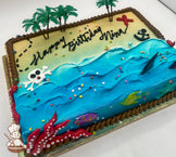 Sheet cake with buttercream decorations featuring a beach and ocean theme, with plastic palm trees, a red "X," an anchor, a skeleton head, plastic shark fins, fish, and a starfish. Rope trim borders the cake.