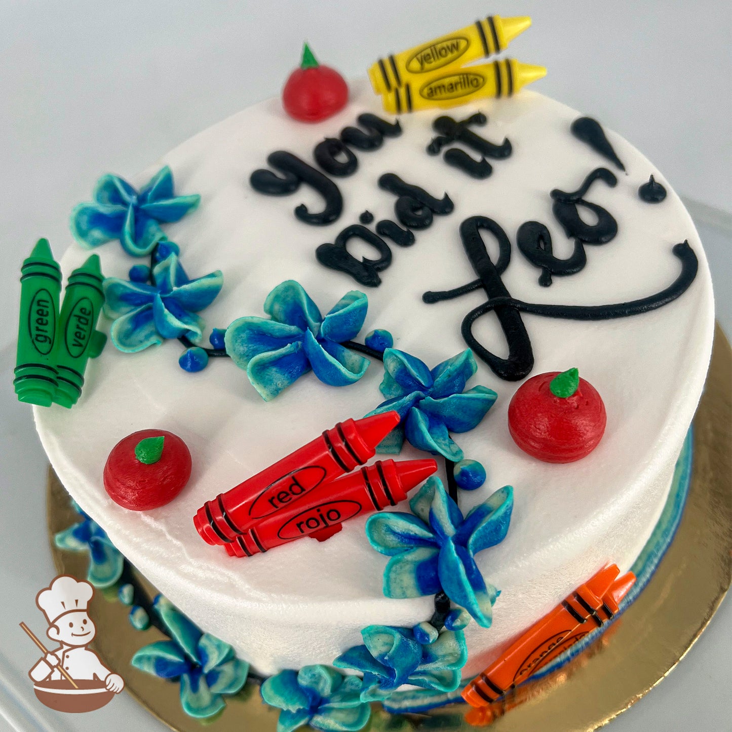 Round white cake decorated with blue buttercream flowers, red apple toppers, and plastic crayons in green, red, yellow, and orange.