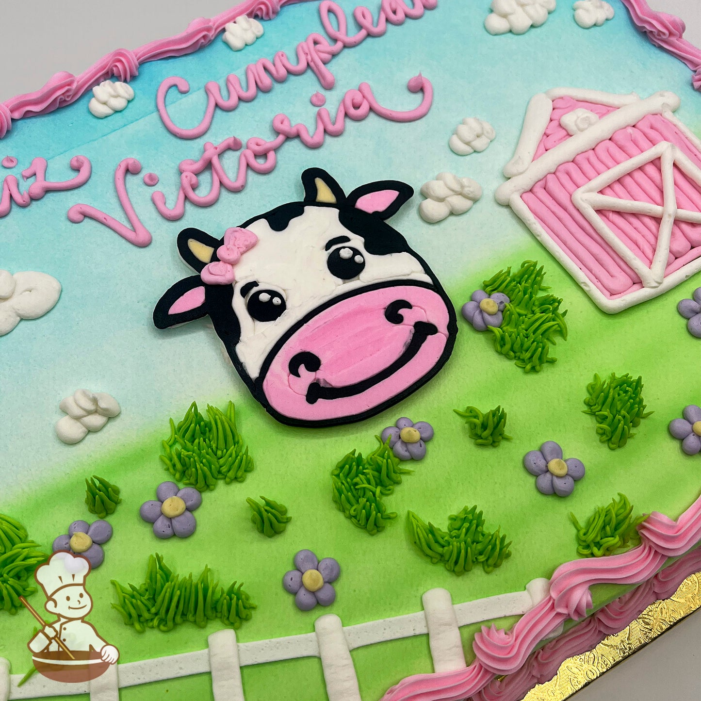 Sheet cake with a buttercream design featuring a smiling cow face, a pink barn, green grass, purple flowers, white clouds, a fence, and a blue sky.