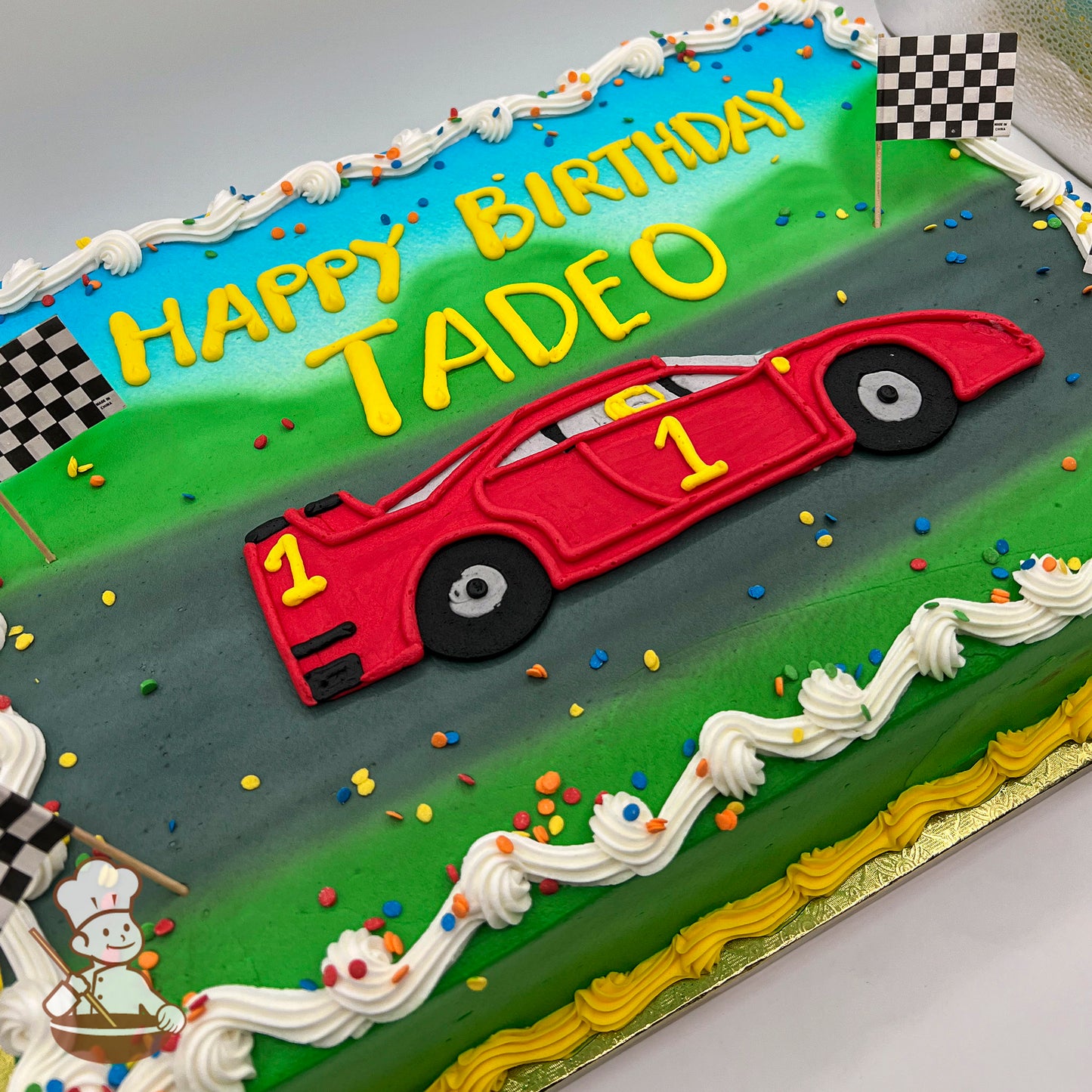 Sheet cake with buttercream decorations featuring a red race car on a gray track, green grass, checkered flags, colorful sprinkles, and white frosting borders.