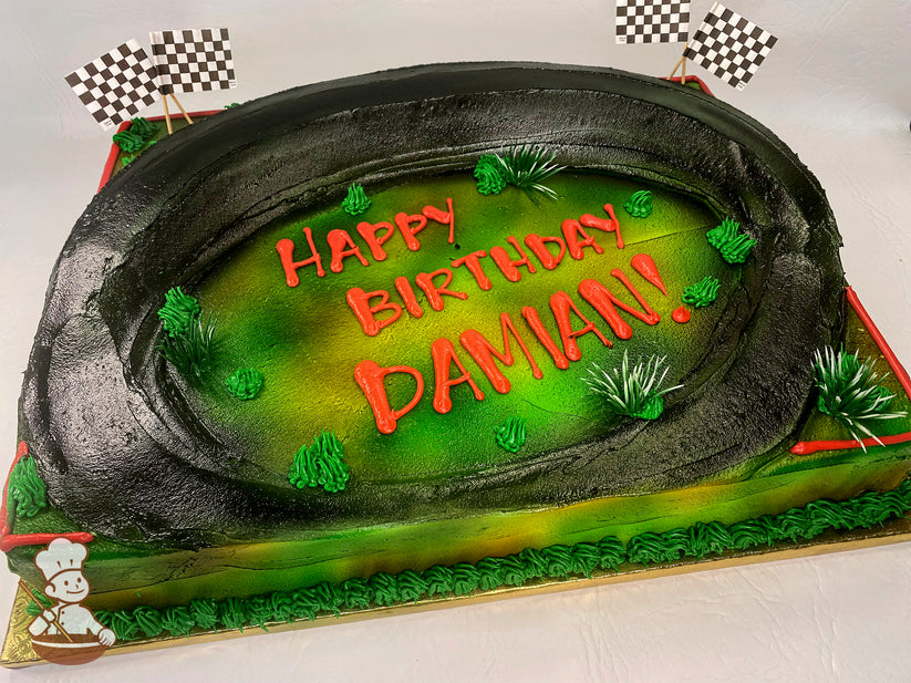 Sheet cake with a black buttercream racetrack, green grass accents, and checkered flag toppers at the corners. Decorated with vibrant colors.