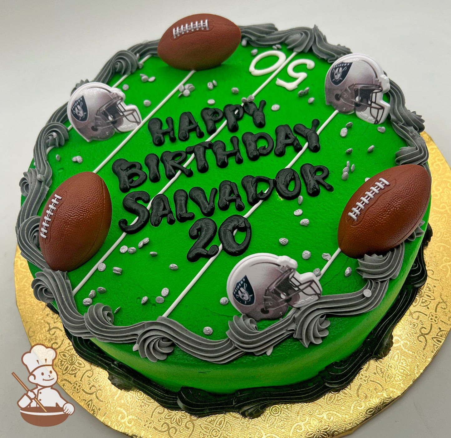Round cake with green icing resembling a football field, decorated with plastic footballs and Raiders helmets, bordered by gray and black buttercream swirls.