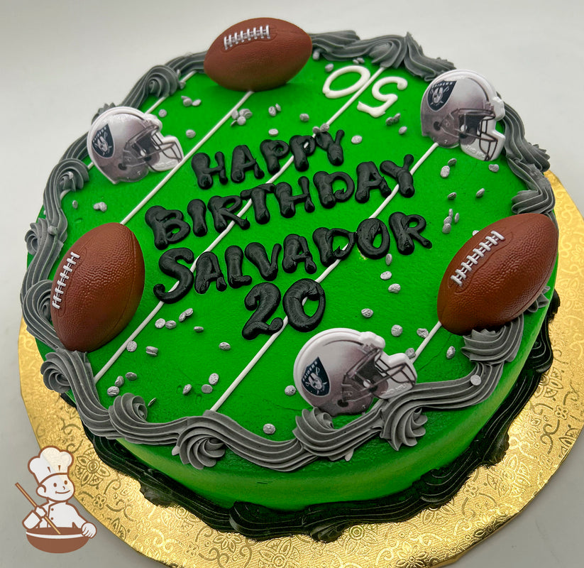 Round cake with green icing resembling a football field, decorated with plastic footballs and Raiders helmets, bordered by gray and black buttercream swirls.