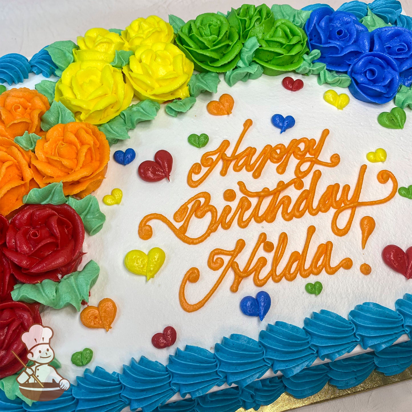 Sheet cake with colorful buttercream roses forming a rainbow arch, surrounded by small heart decorations in various colors, and a blue frosting border.
