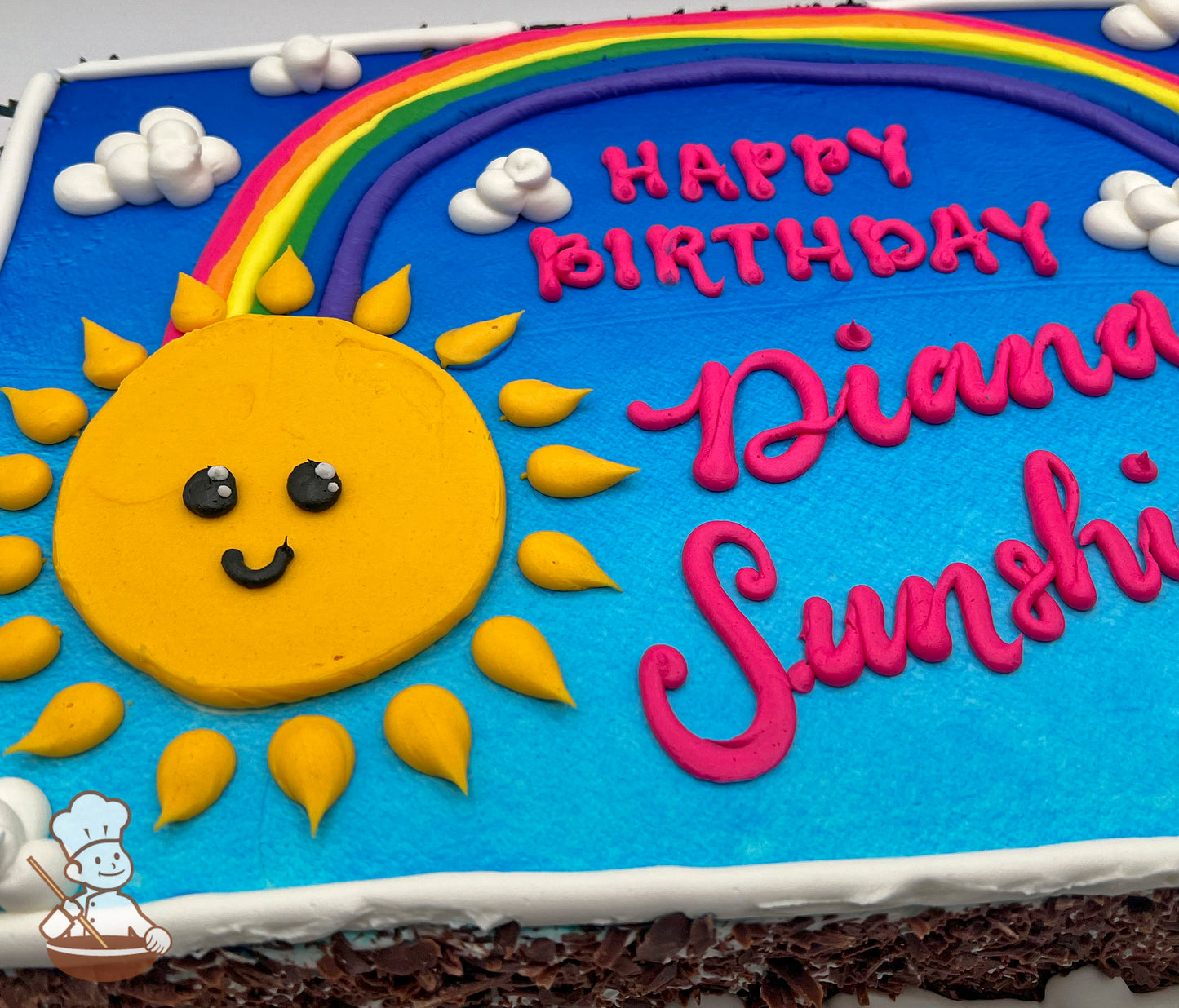 A sheet cake with blue frosting, decorated with a smiling yellow sun, a colorful rainbow, and white buttercream clouds.