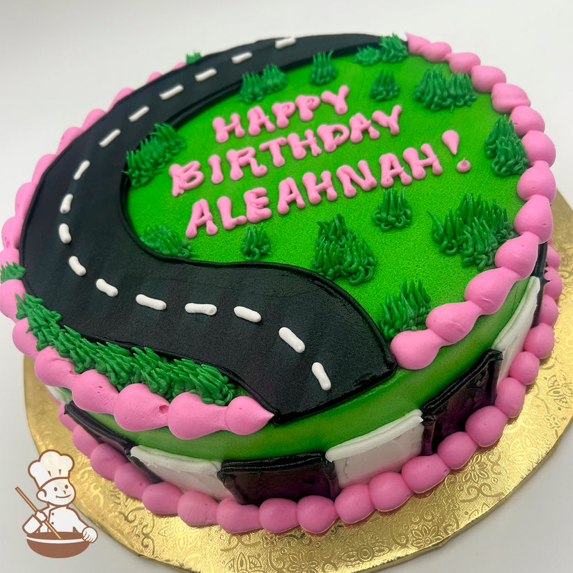 Round sheet cake with a black buttercream road curving across green grass-like frosting, pink border, and small grass tufts.