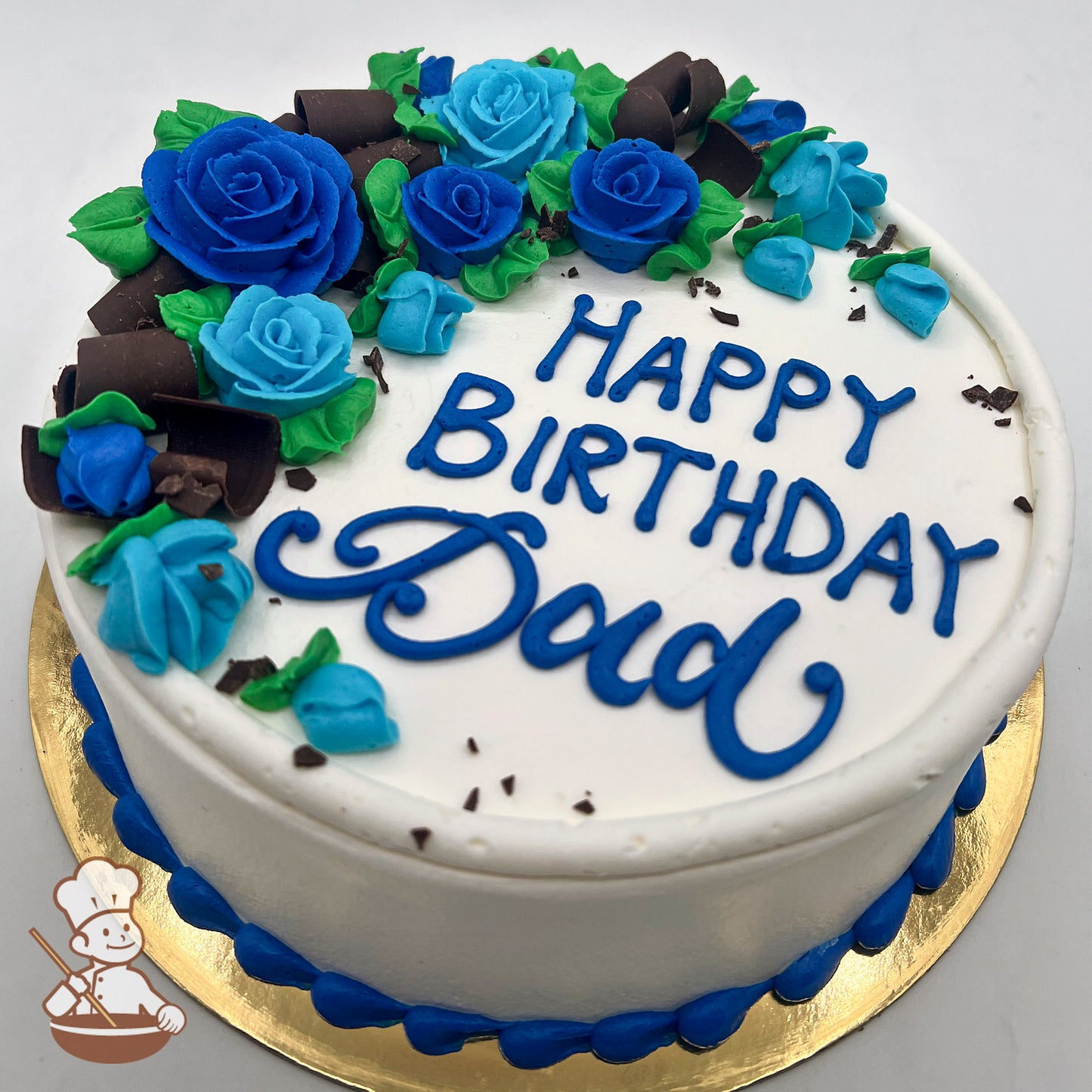 Round white cake decorated with blue buttercream roses, green leaves, and chocolate curls on top, with a blue border at the base.