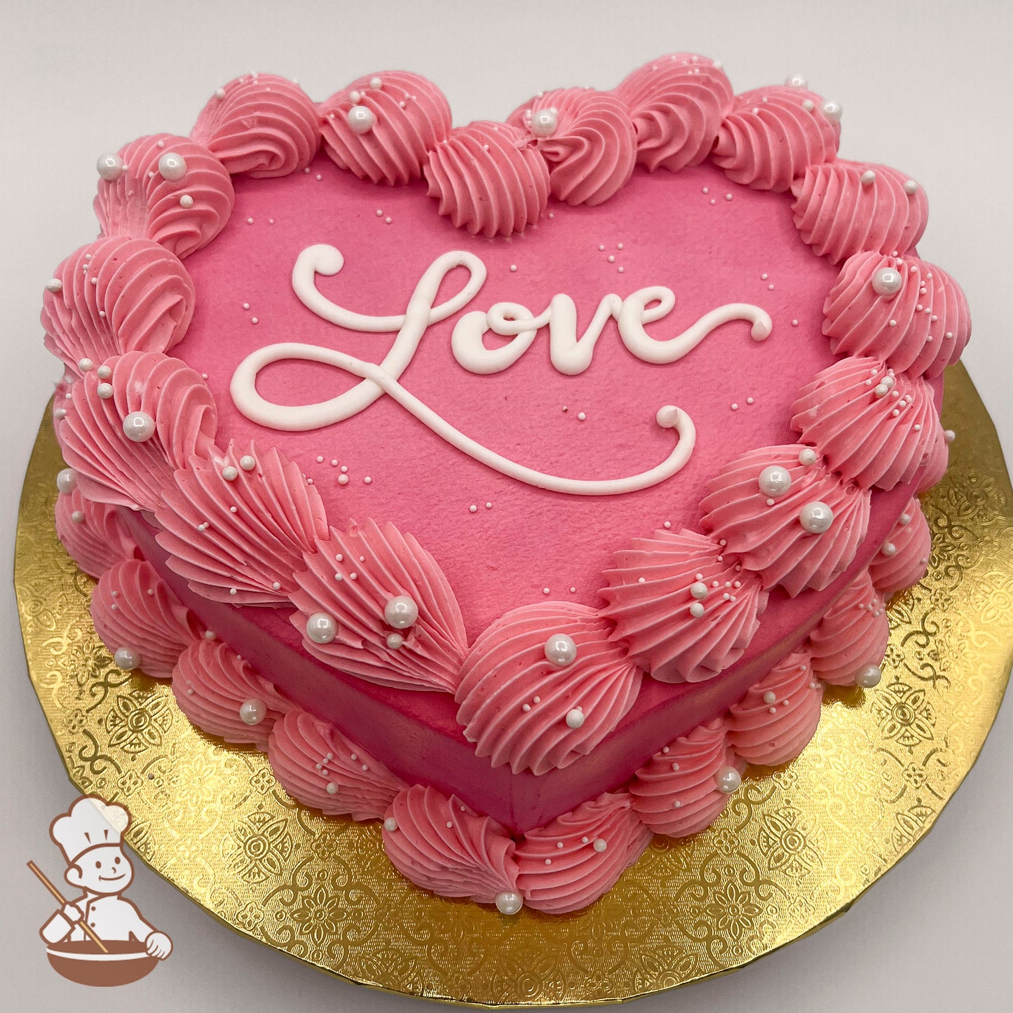 Heart-shaped cake with pink icing, scallop trim, and sugar pearls on a gold base.