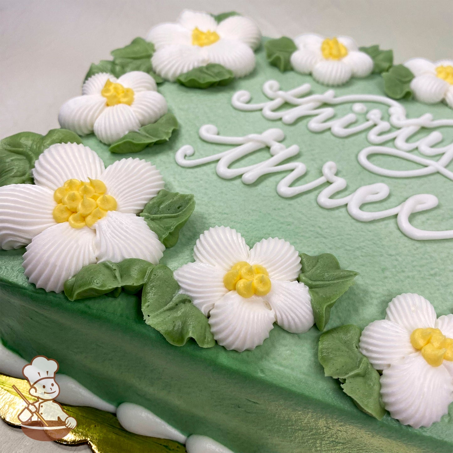 Light green sheet cake with white buttercream daisies featuring yellow centers, surrounded by green buttercream leaves. White piped border on the base.