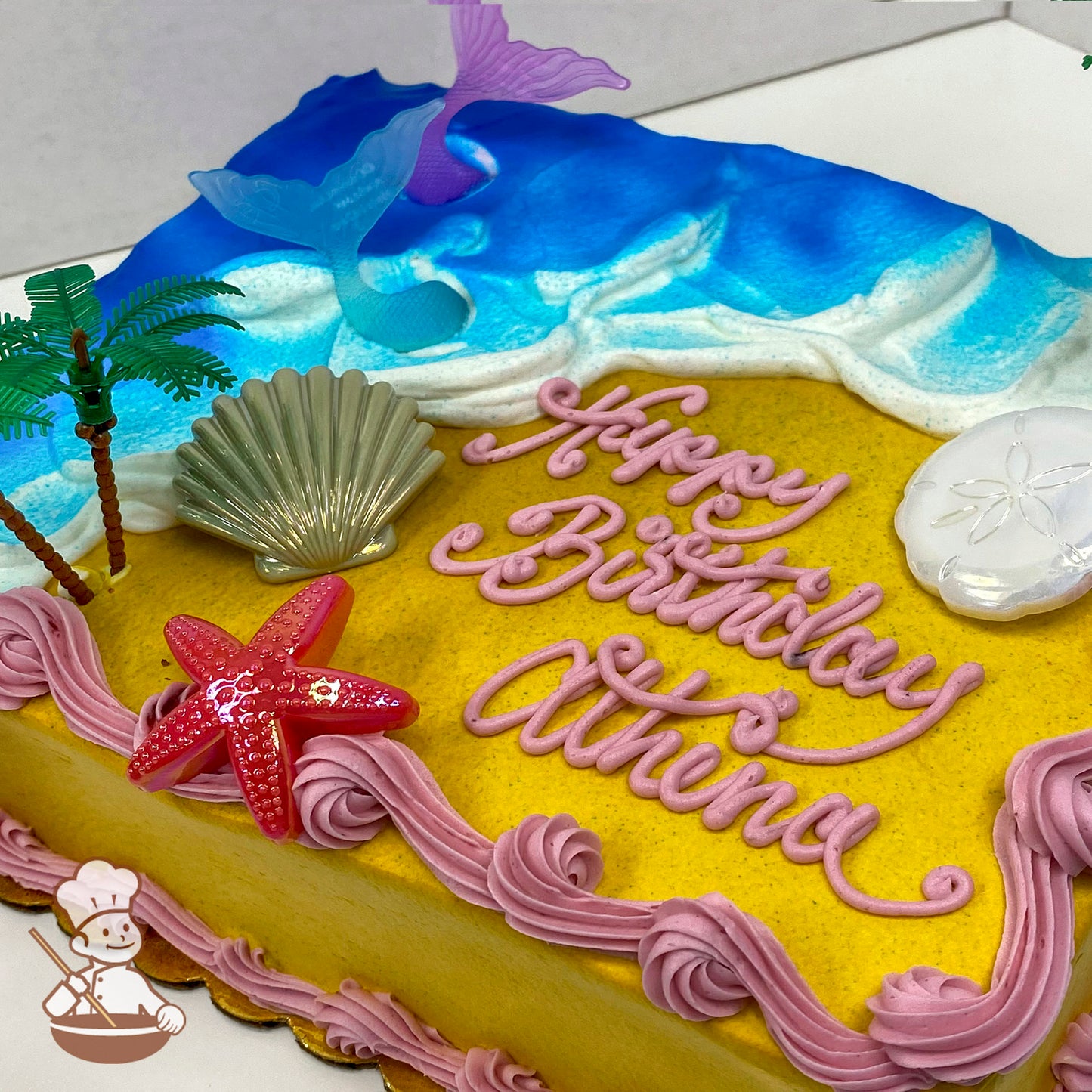 Sheet cake with a beach and mermaid theme, featuring buttercream waves, sand, plastic palm trees, mermaid tails, a starfish, a seashell, and a sand dollar.
