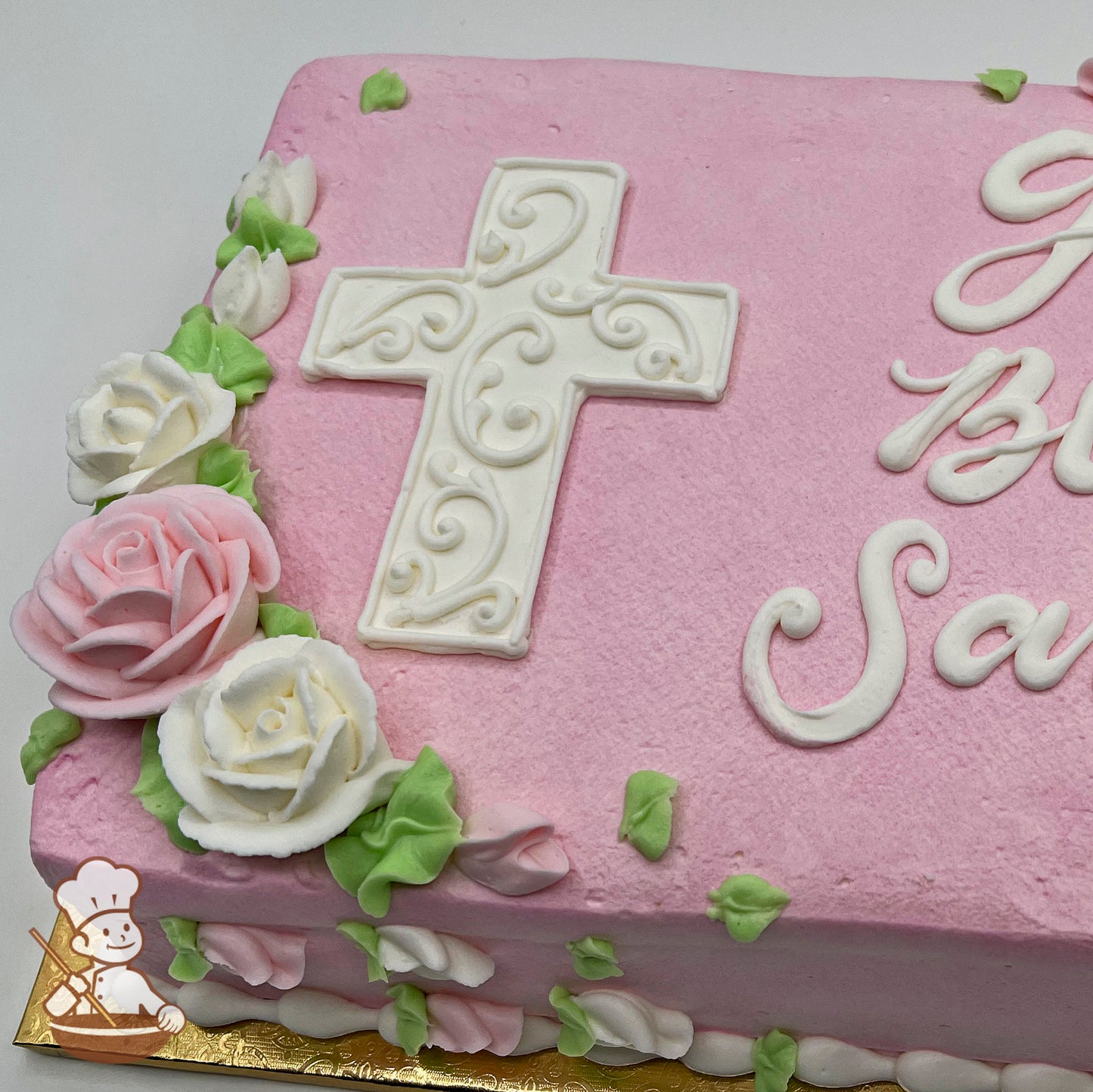 Pink sheet cake with a white buttercream cross featuring intricate scroll designs, surrounded by pink and white buttercream roses and green leaves.