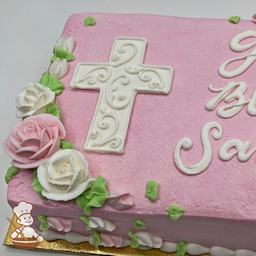 Pink sheet cake with a white buttercream cross featuring intricate scroll designs, surrounded by pink and white buttercream roses and green leaves.