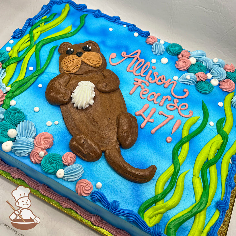 Sheet cake with a blue base, featuring a brown buttercream sea otter holding a white shell, surrounded by green kelp, pink and blue swirls, and white bubbles.