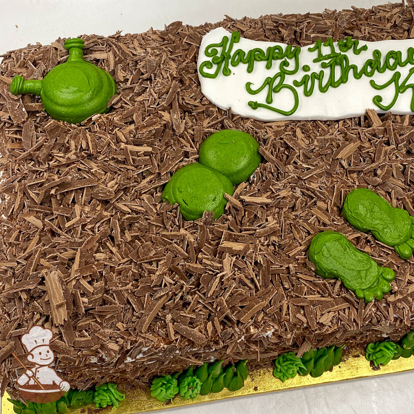 Sheet cake decorated with Belgian chocolate shavings resembling mud, green buttercream Shrek figure partially submerged, and Shrek's footprints.
