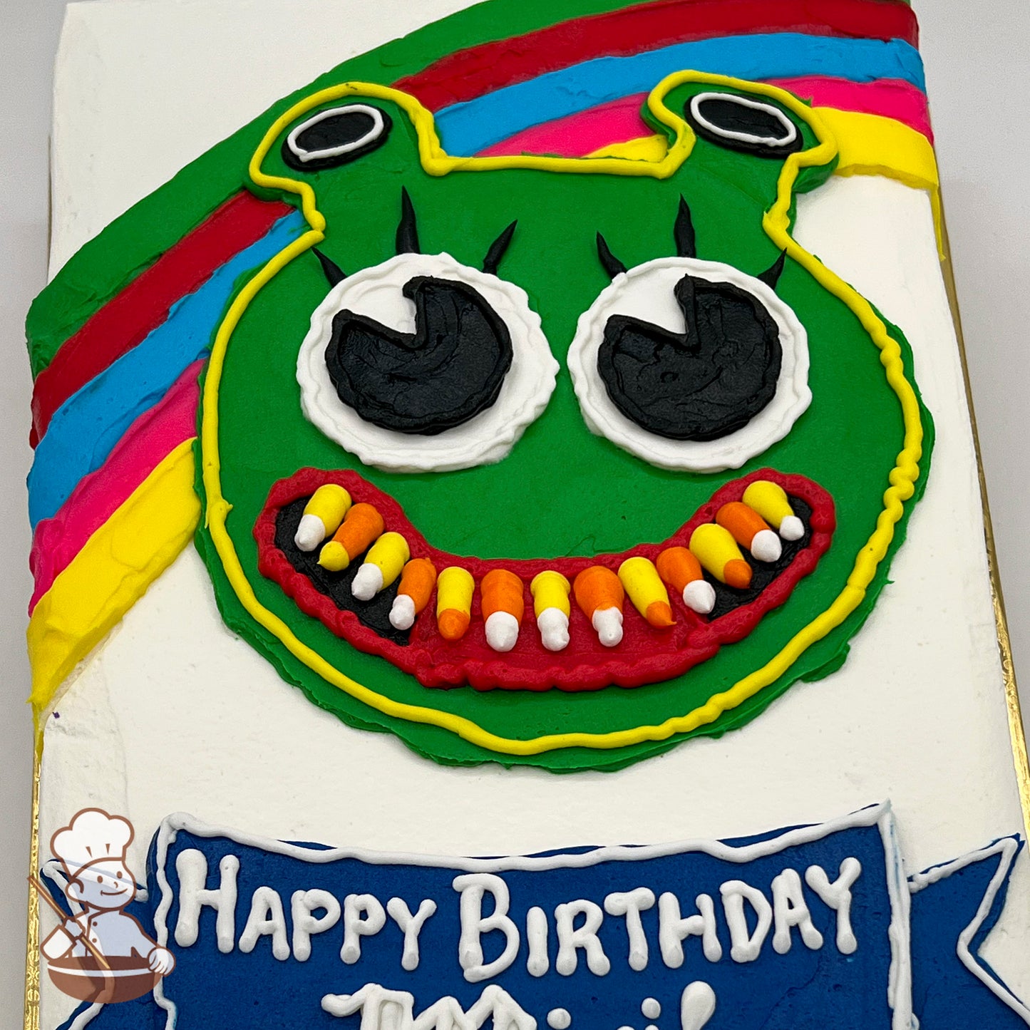 A sheet cake with a green ogre face made of buttercream, featuring large black and white eyes, candy corn teeth, and a colorful rainbow background.