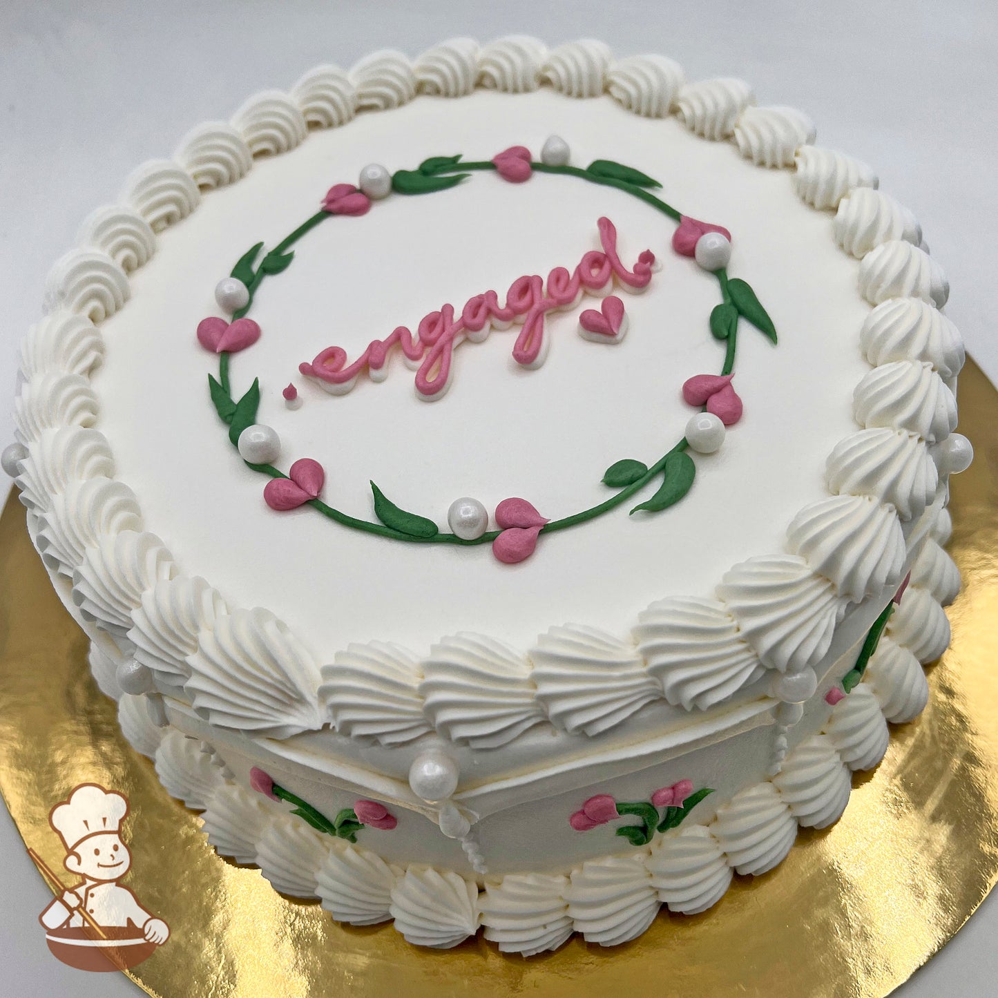 Round white cake, decorated with pink heart flower buds, green leaves, sugar pearls, and piped white frosting borders, on a gold base.