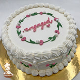 Round white cake, decorated with pink heart flower buds, green leaves, sugar pearls, and piped white frosting borders, on a gold base.
