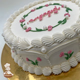 Round white cake, decorated with pink heart flower buds, green leaves, sugar pearls, and piped white frosting borders, on a gold base.
