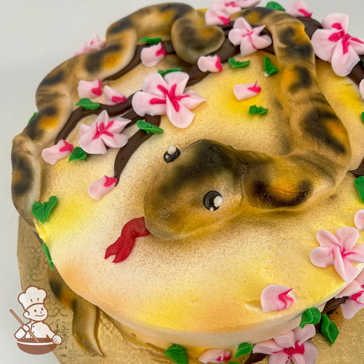 A sheet cake decorated with a realistic buttercream snake, coiled on a yellow surface, surrounded by pink cherry blossoms and green leaves.