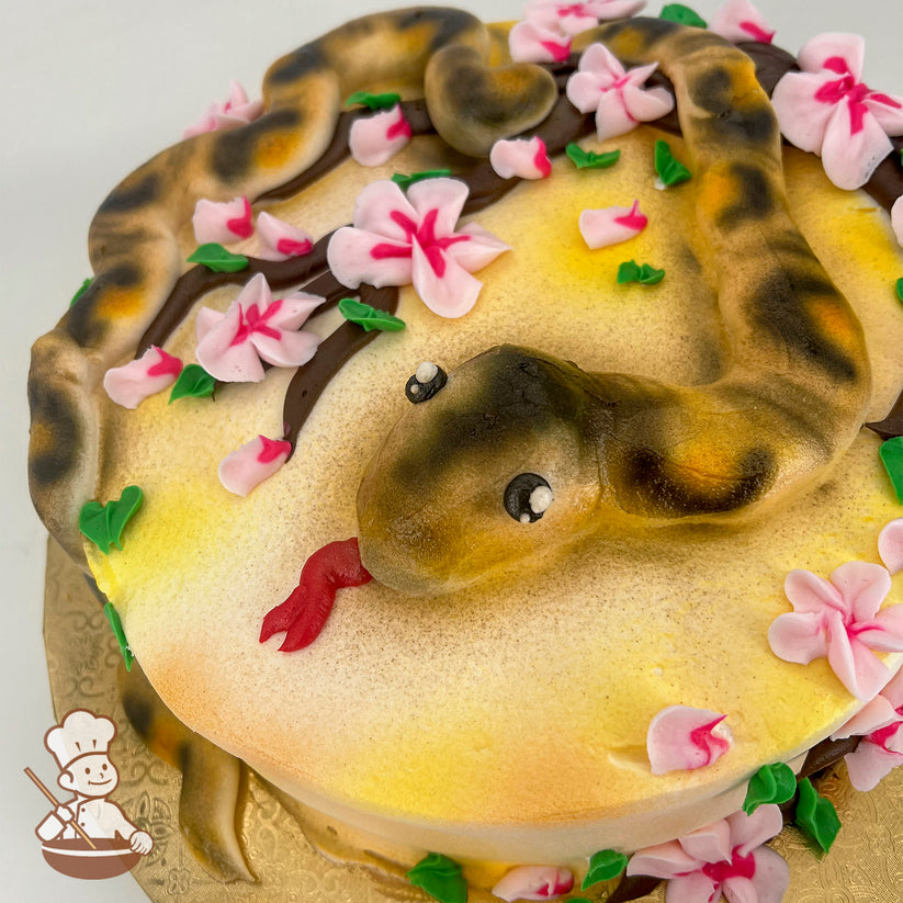 A sheet cake decorated with a realistic buttercream snake, coiled on a yellow surface, surrounded by pink cherry blossoms and green leaves.