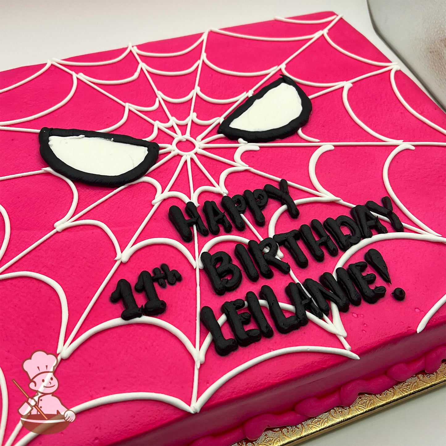 Pink sheet cake with white spider web design, black-outlined white Spider-Girl eyes, and piped black buttercream text.