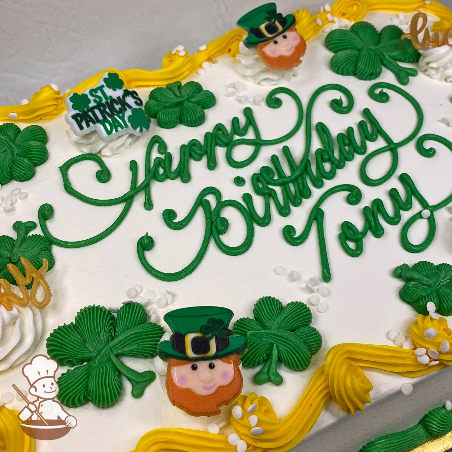 Sheet cake with green buttercream shamrocks, a leprechaun face, and yellow swirls. Plastic St. Patrick's Day sign and white sprinkles.