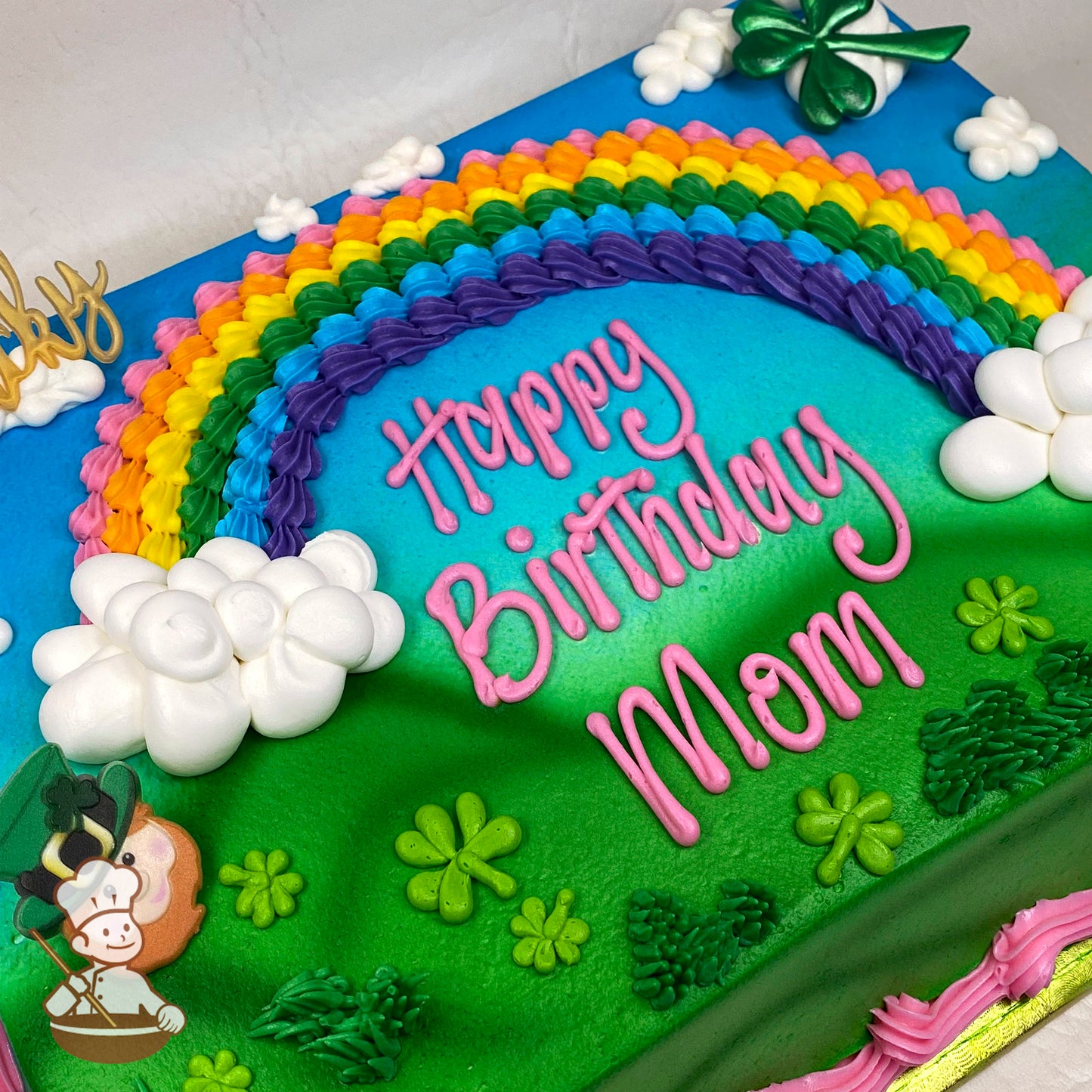 Sheet cake with a colorful buttercream rainbow, white clouds, green clovers, grass details, and a leprechaun plastic face decoration.