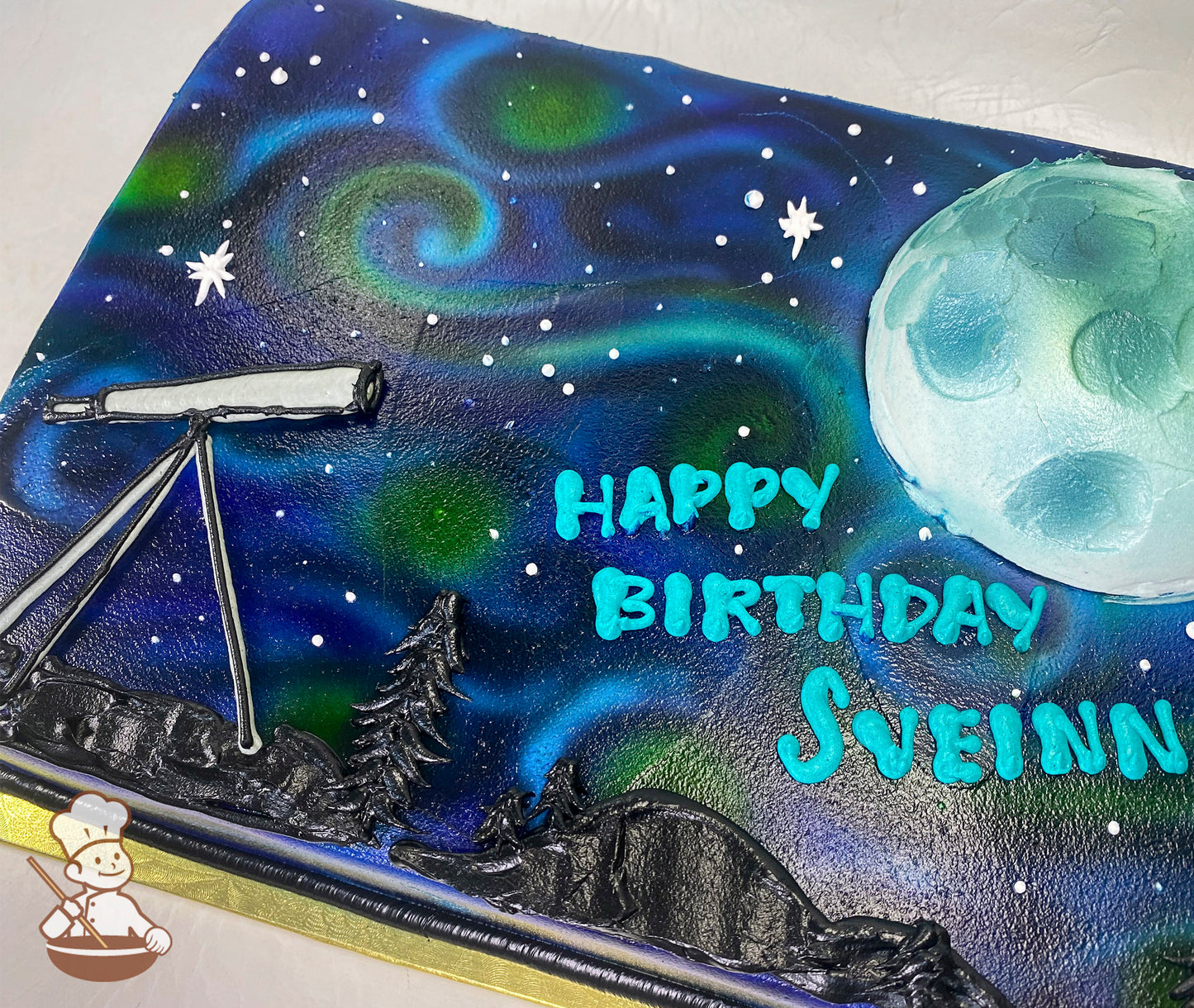 Sheet cake with a night sky theme, featuring a buttercream telescope, moon, stars, trees, and mountains. Swirling blue and green sky.