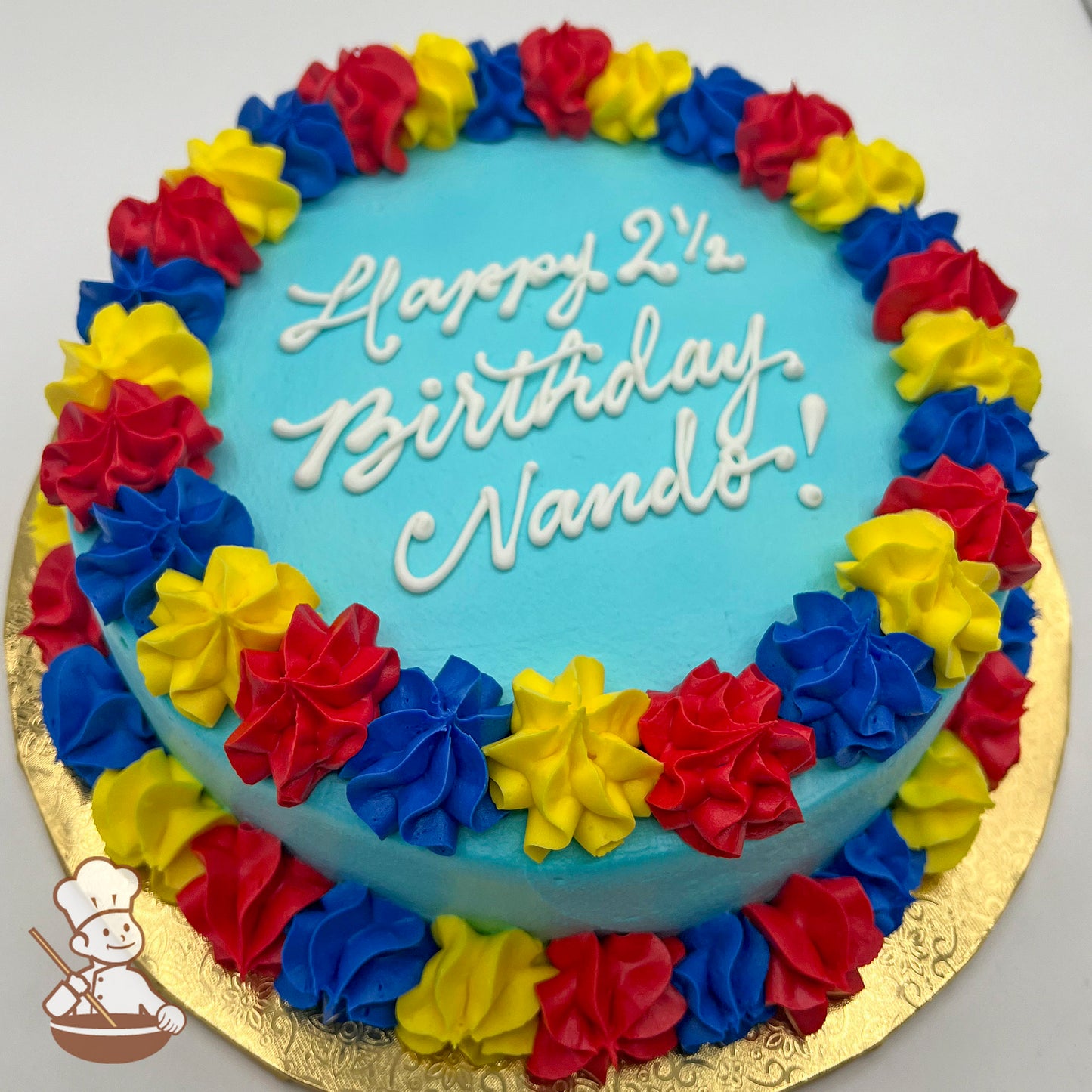 Round sheet cake with blue frosting, decorated with a border of red, yellow, and blue star-tip buttercream swirls on a gold base.