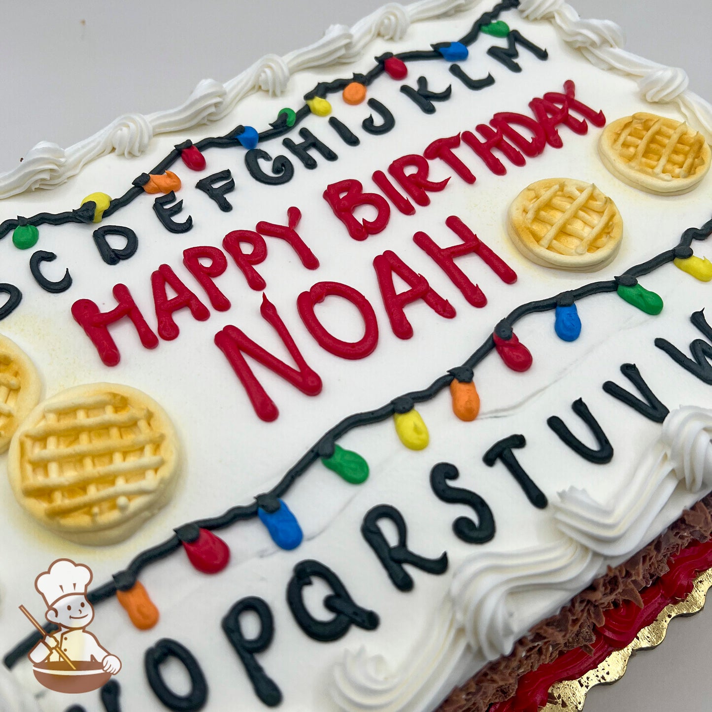 Sheet cake with white frosting, decorated with colorful string lights, black alphabet letters, and waffle designs.