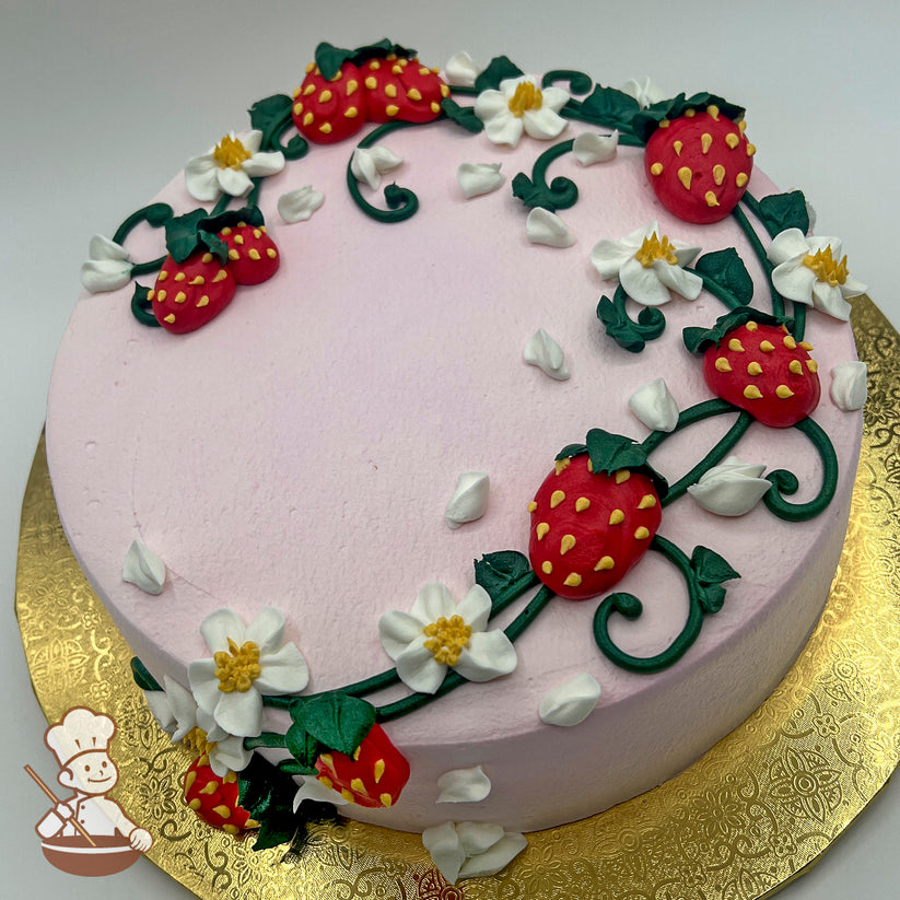 Round pink sheet cake with buttercream decorations of strawberries, white blossoms, green vines, and scattered petals on a gold base.