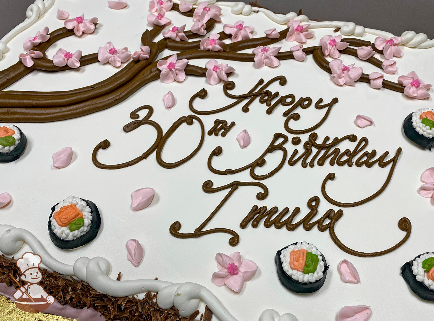 Sheet cake with buttercream cherry blossom tree and sushi decorations. Pink cherry blossoms and sushi rolls are scattered across the white icing.