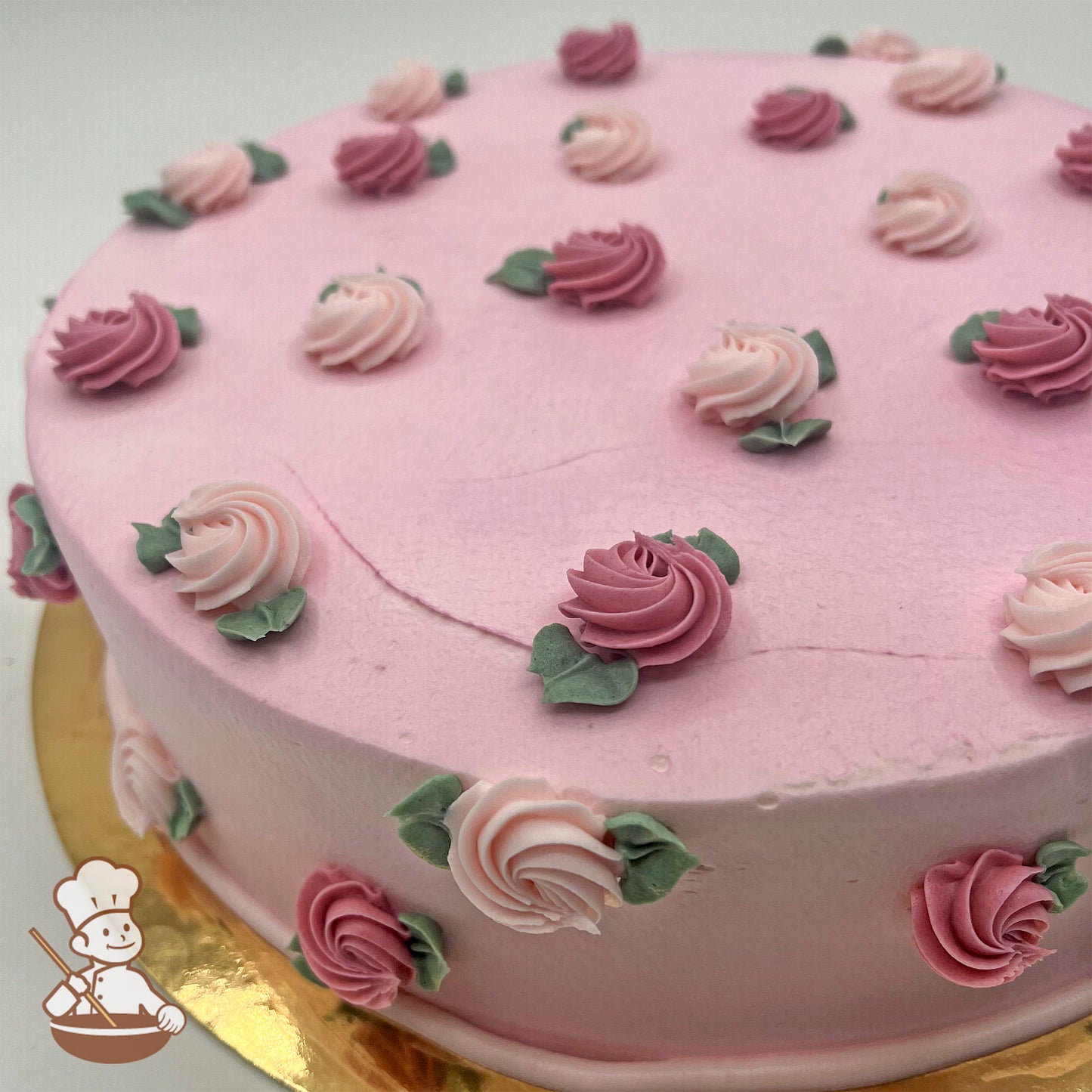 Pink cake decorated with swirled rosette buds in light pink and dark pink, accented with small green leaves, on a gold cake board.