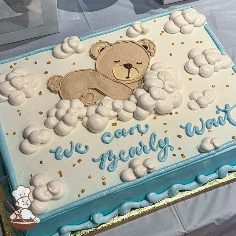 A sheet cake with a light blue border and white frosting. It features a sleeping teddy bear design surrounded by fluffy white buttercream clouds and gold sprinkles.
