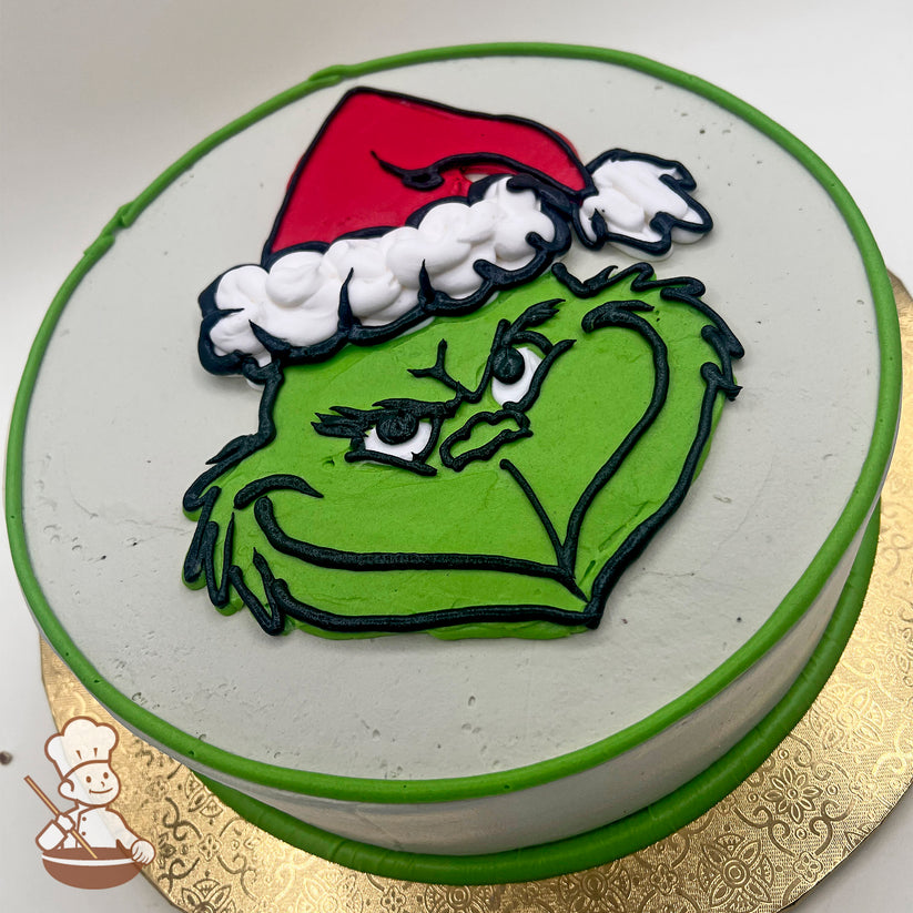 Round sheet cake with a buttercream design of the Grinch's green face wearing a red Santa hat, outlined in black, on a white background.