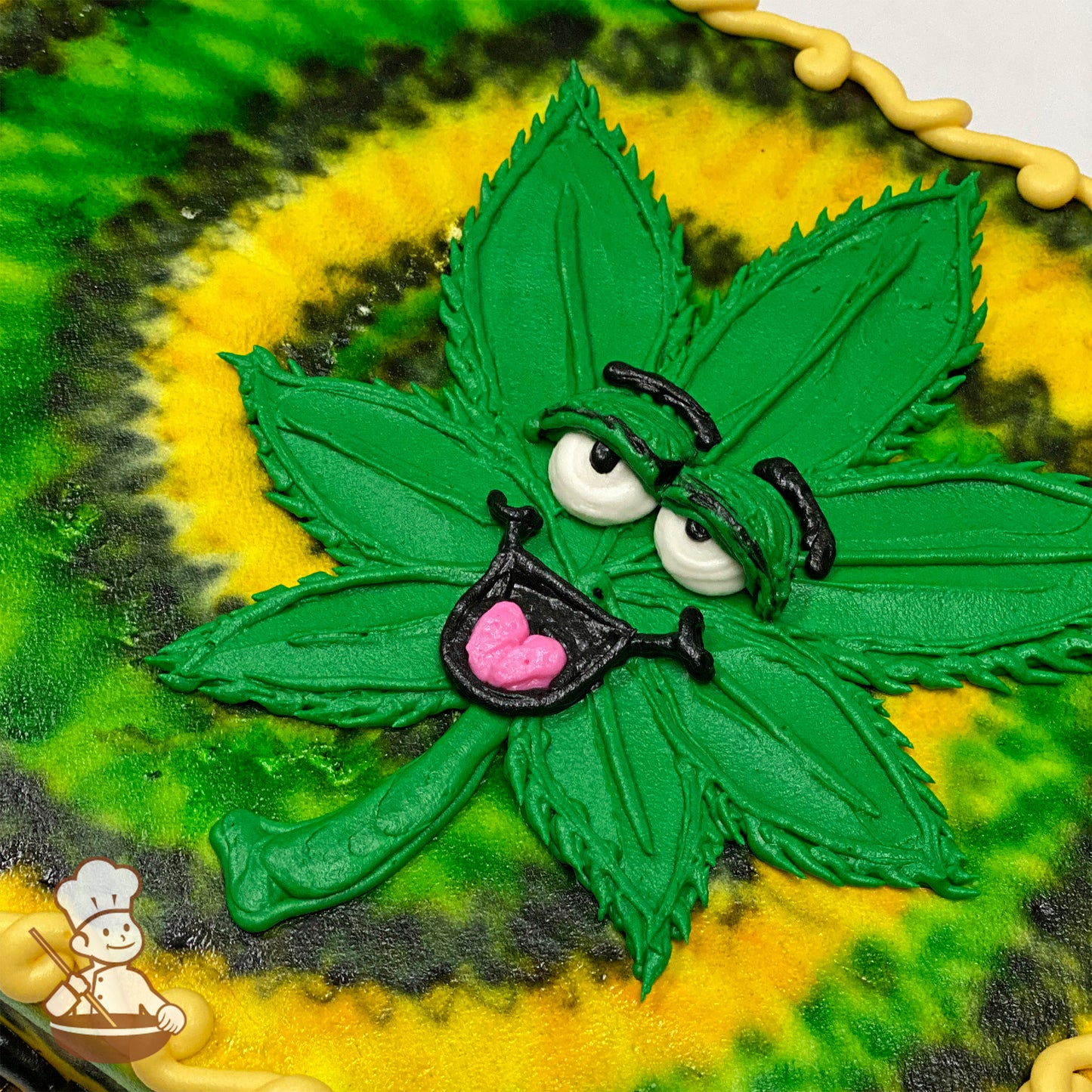 A sheet cake with a green marijuana leaf made of buttercream, featuring a happy face with expressive eyes and a pink tongue. Swirl background in yellow and green.