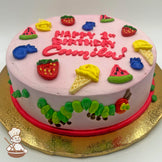 Round cake with pink icing, decorated with colorful fruits, ice cream cones, and a caterpillar design around the side.